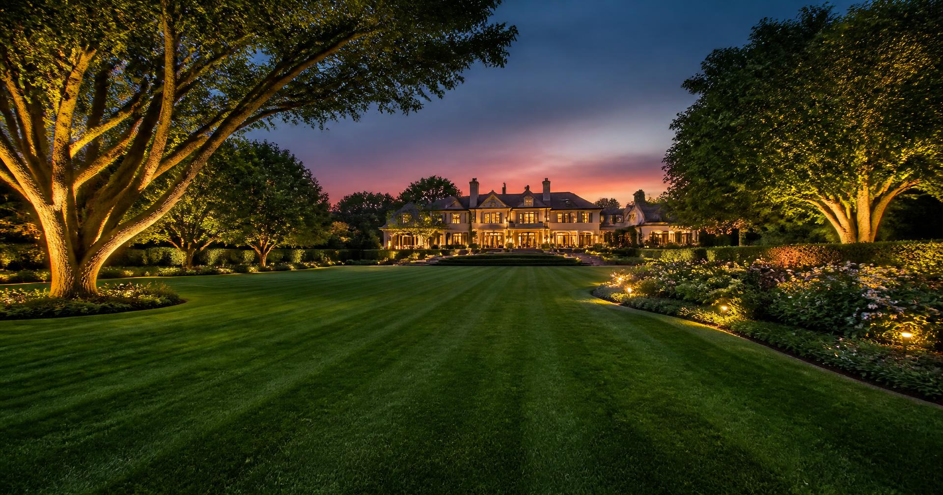 Expansive manicured estate lawn with mature trees and landscape lighting at twilight